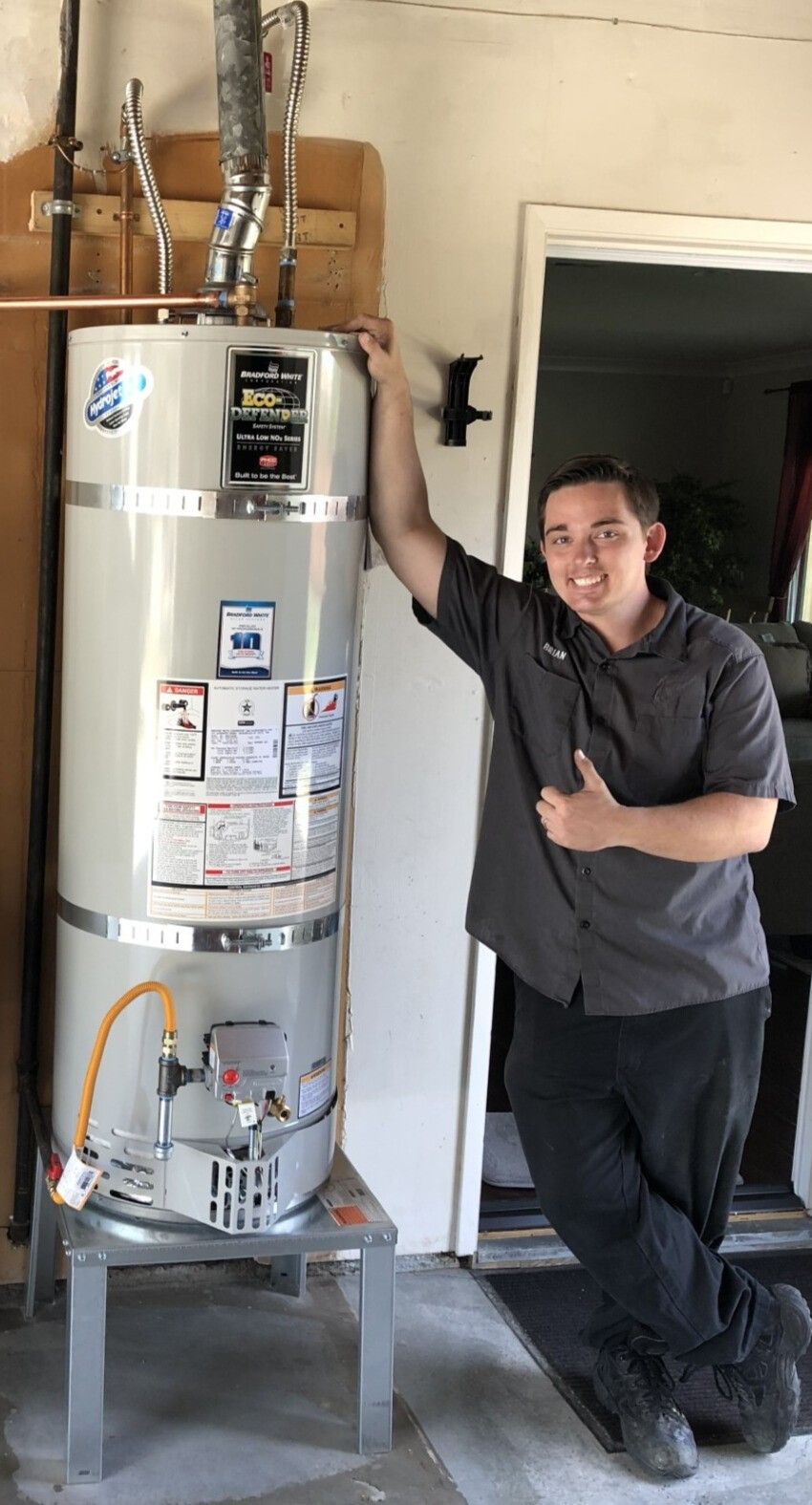 Man stands next to a tall water heater. Gray and silver, inside a garage, smiling.
