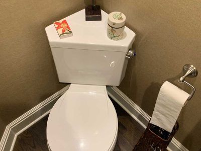 White corner toilet in a small bathroom, with items on the tank lid, and a roll of toilet paper to the right.