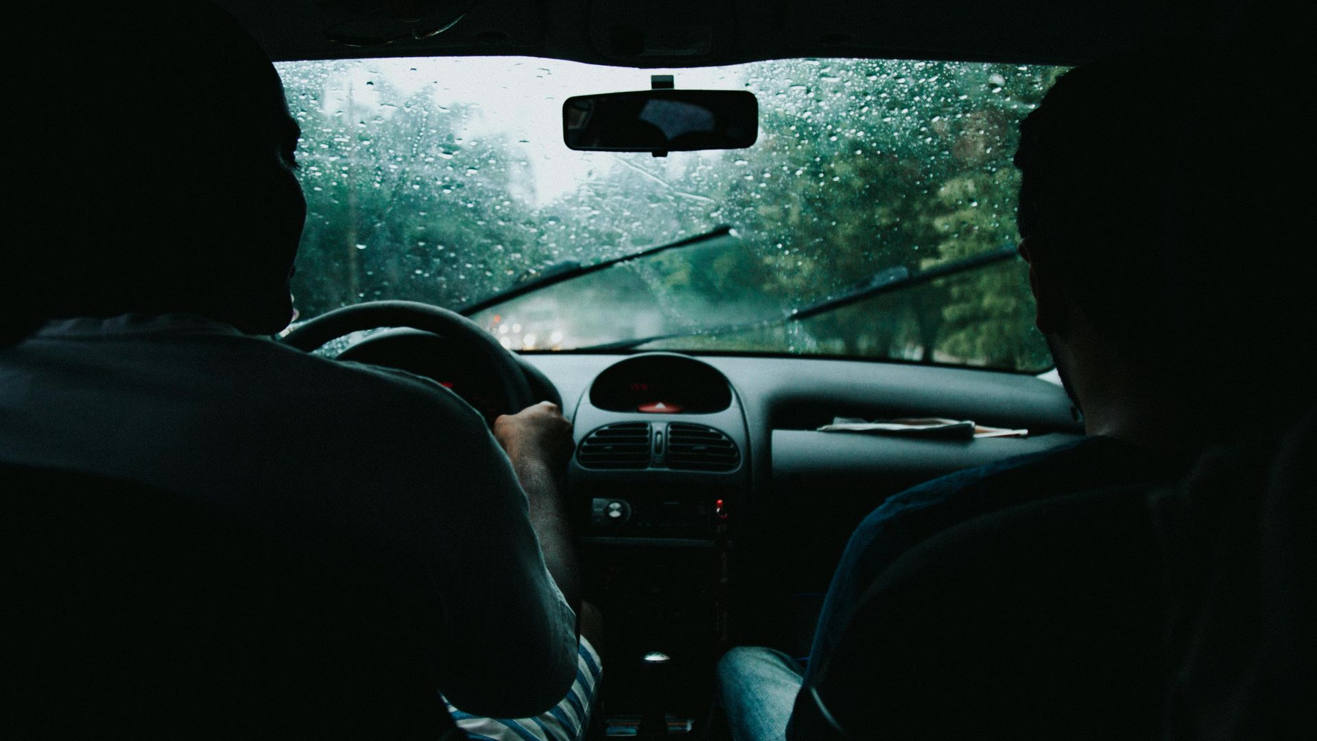 Two people in a car driving in heavy rain; windshield wipers active, trees visible outside.