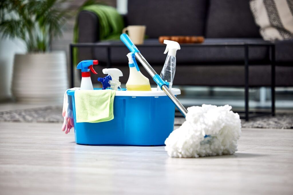 Cleaning supplies in a blue bucket with a mop on a hardwood floor, in a living room setting