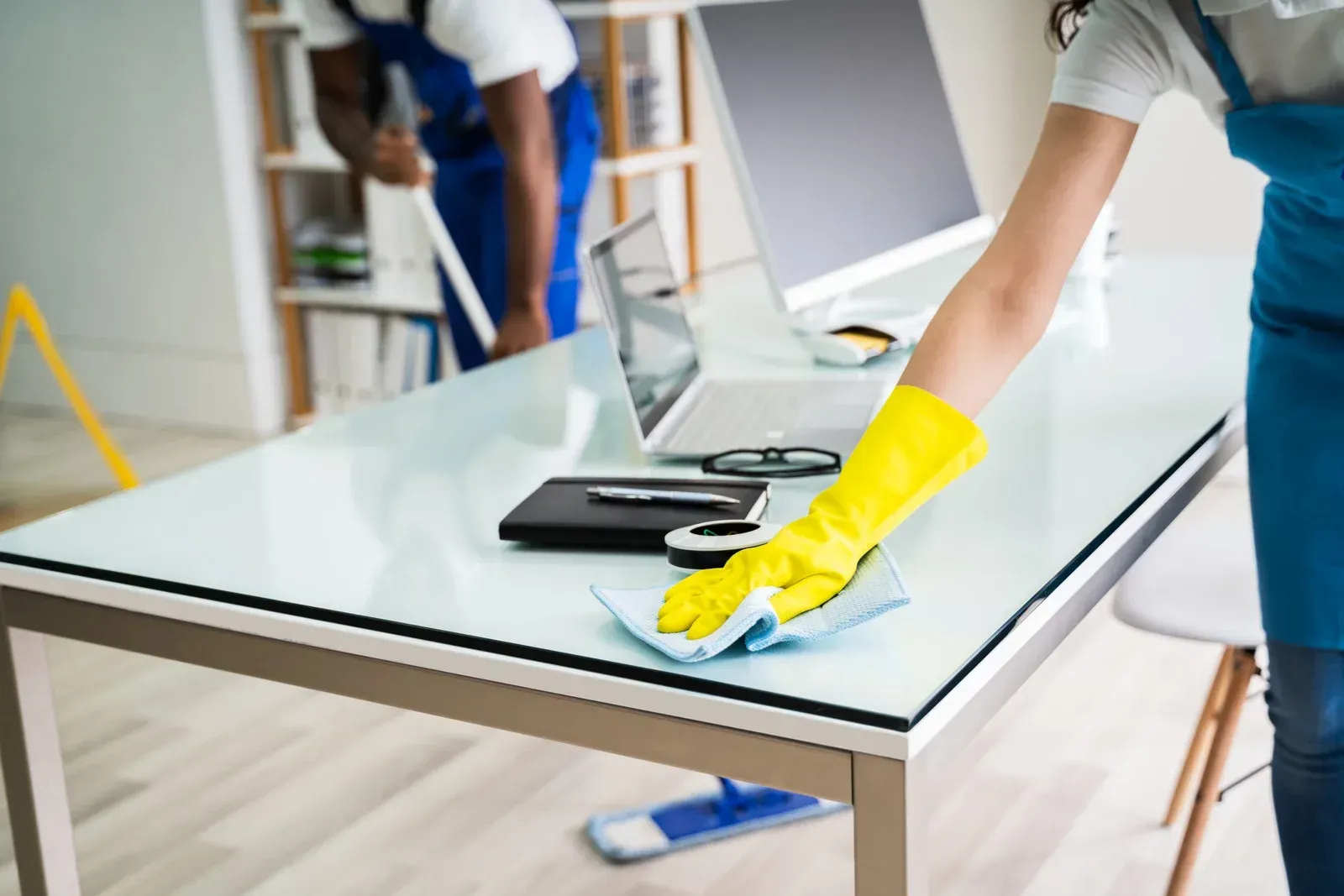 People cleaning a glass table with yellow gloves and spray in a bright office room