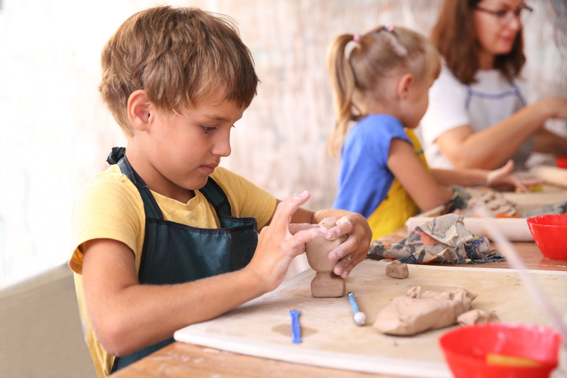 Boy sculpting clay, focused expression, apron, yellow shirt. Other kids and adult in background.