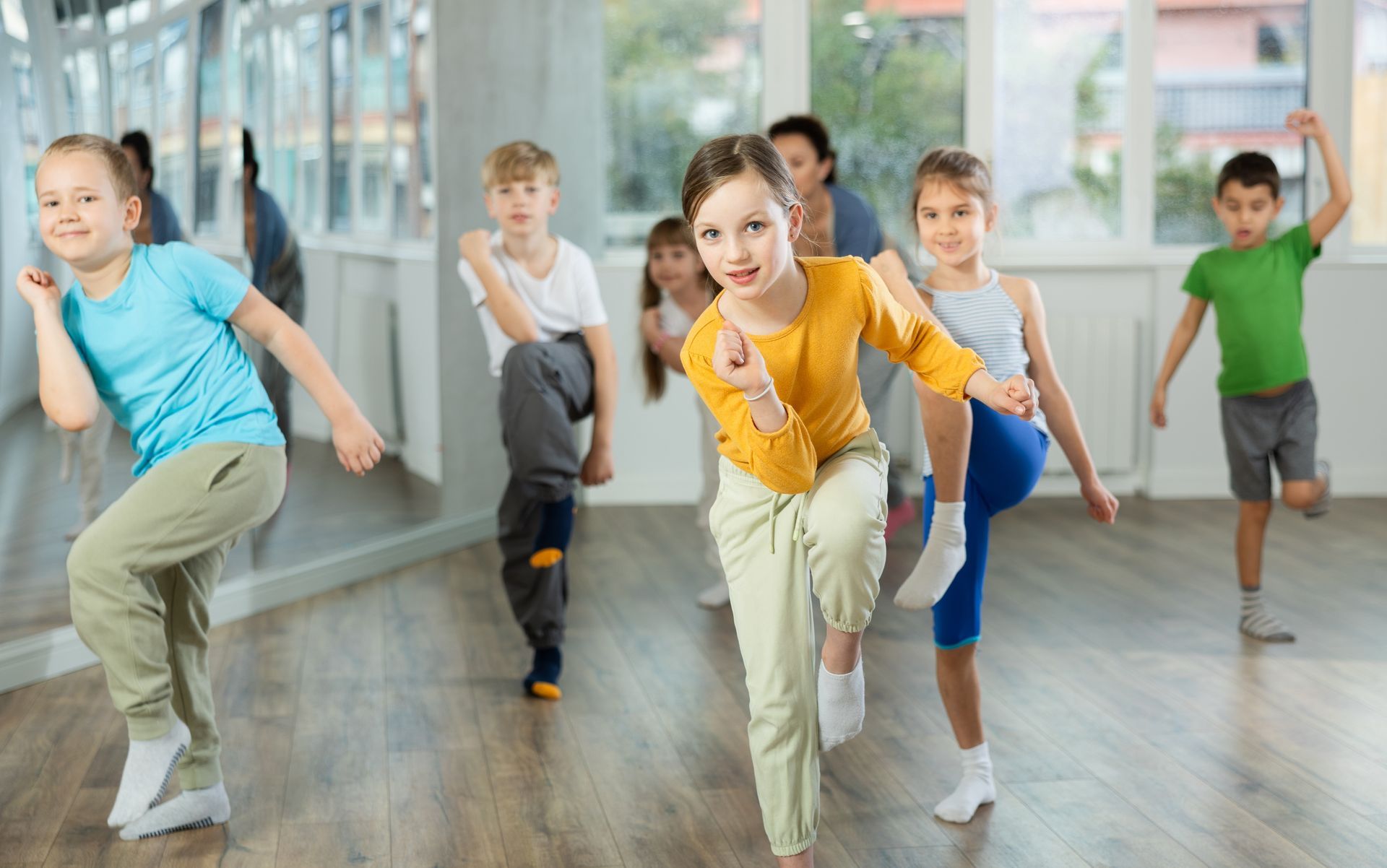 Children dancing in a studio, led by a teacher. Energetic poses, bright colors, wooden floor, and mirrors.