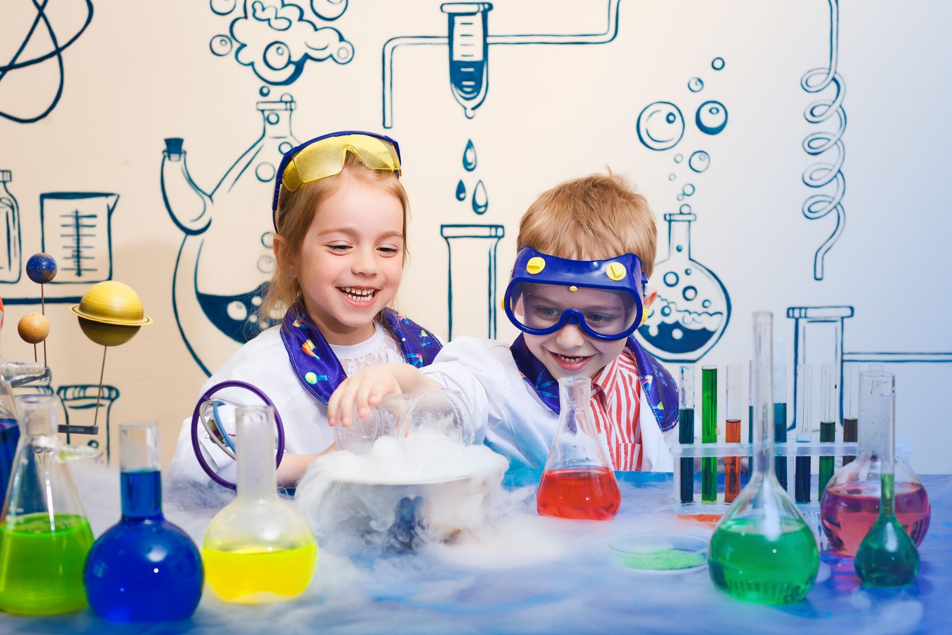 Two children in lab coats and goggles conduct science experiment, with colorful liquids and smoke, smiling.