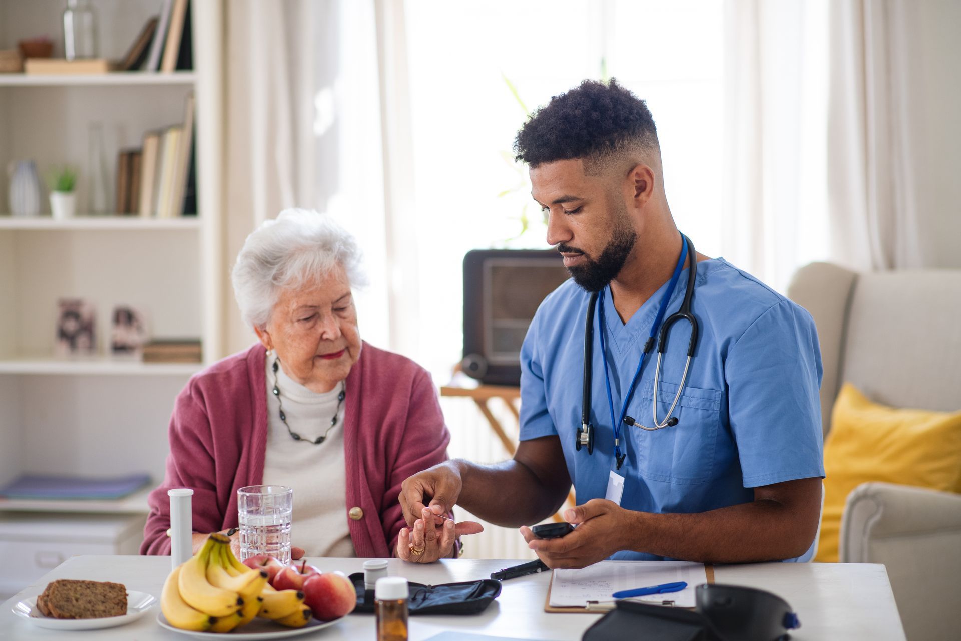 Male nurse checks an elderly woman’s blood sugar at a table. They are indoors with fruit, bread, and a glass of water present.