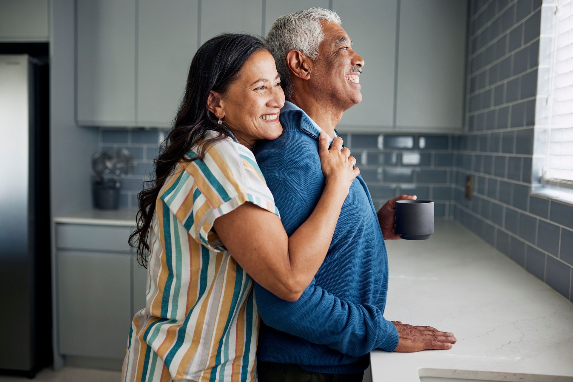 An older couple embraces by a kitchen window. The woman hugs the man from behind, both smiling and looking out.