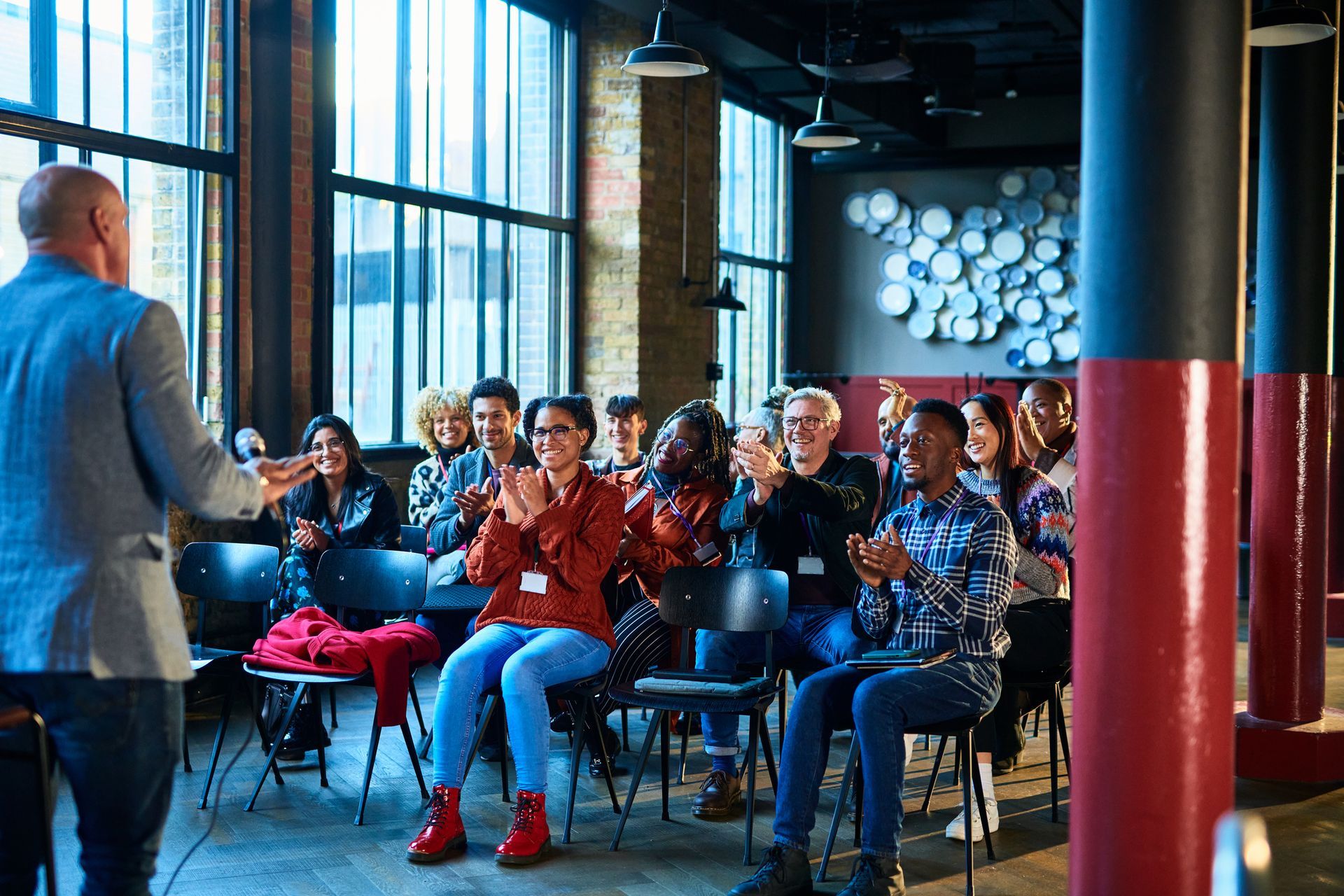 A man lectures to a diverse group clapping in an industrial-chic room with brick and large windows.