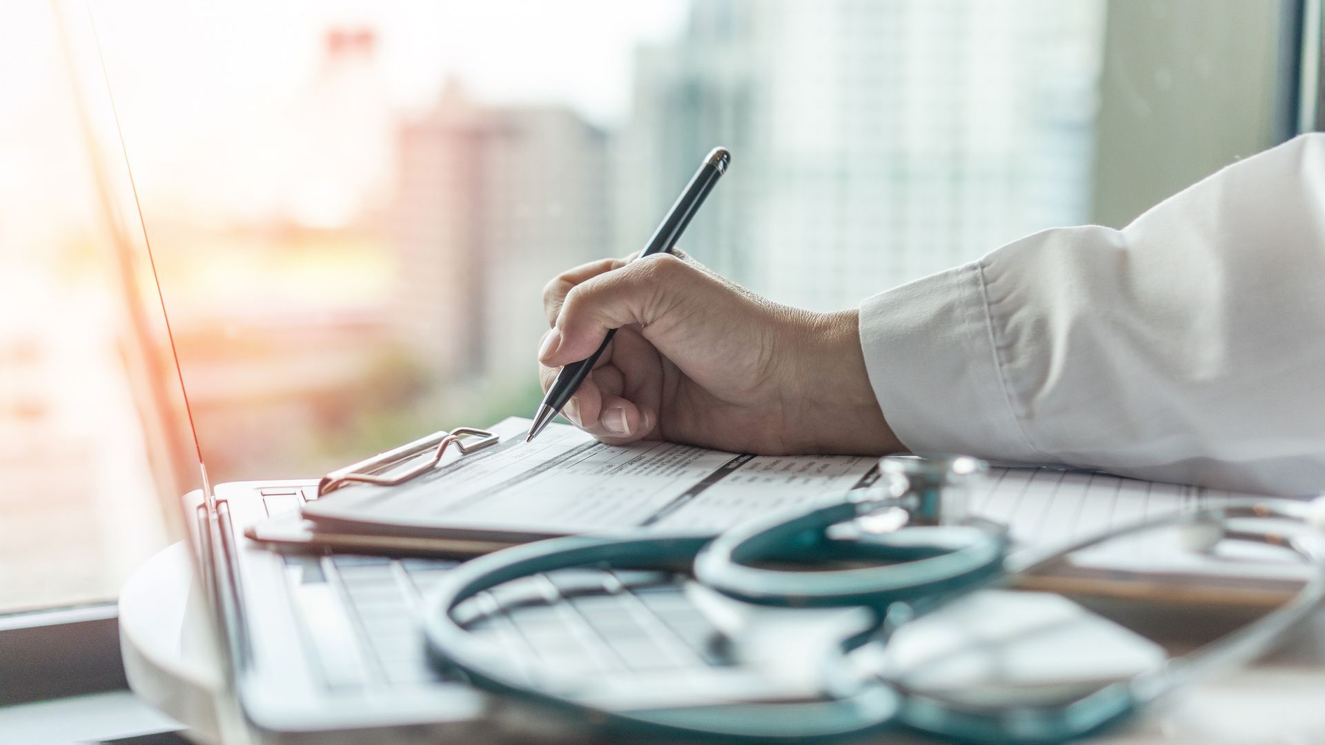 Doctor's hand writing on a clipboard, next to a laptop and stethoscope; bright sunlight streams in from a window.