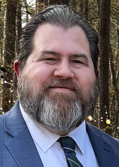 Bearded man in a blue suit and patterned tie smiles outdoors. Background of trees with soft lighting.