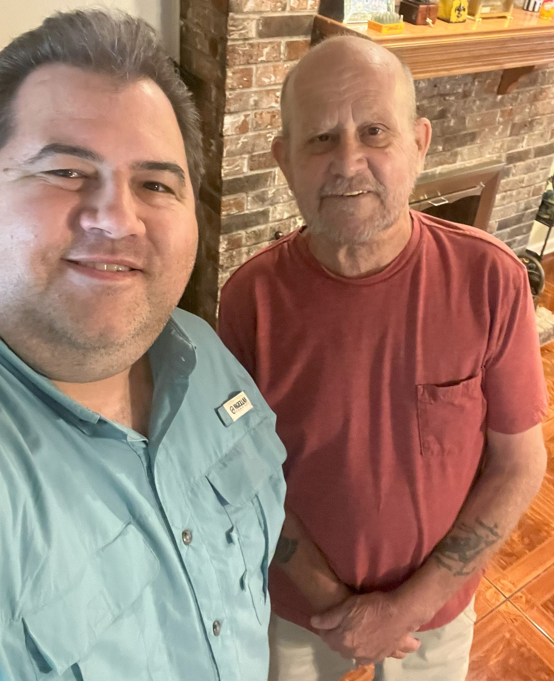 Two men smiling indoors; one in a blue shirt, the other in a red shirt. They stand near a brick wall and fireplace.