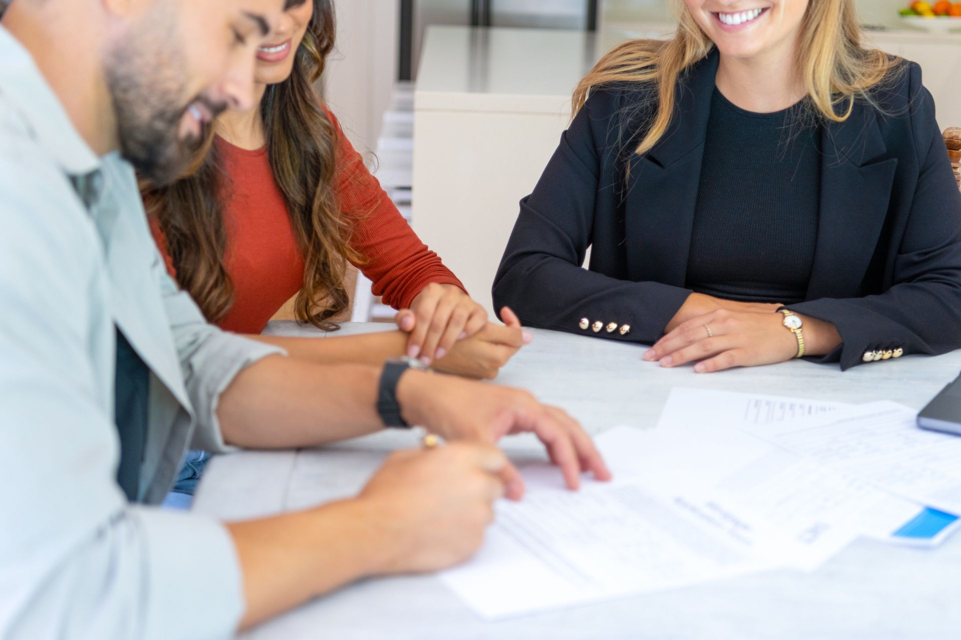 A couple signing documents at a table with a smiling professional in a black blazer, likely a real estate agent.