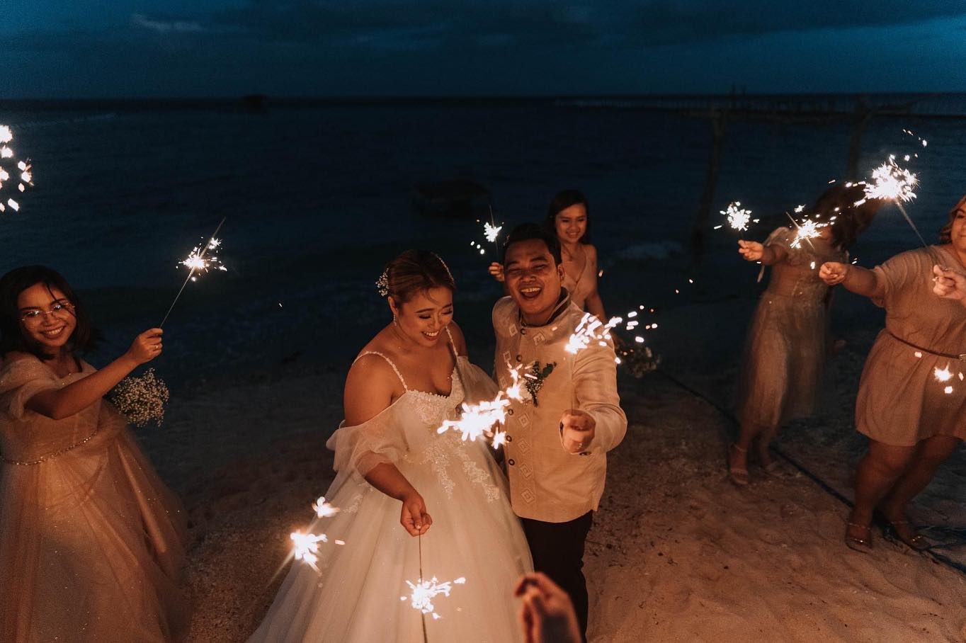 A bride and groom are holding sparklers at their wedding reception on the beach.