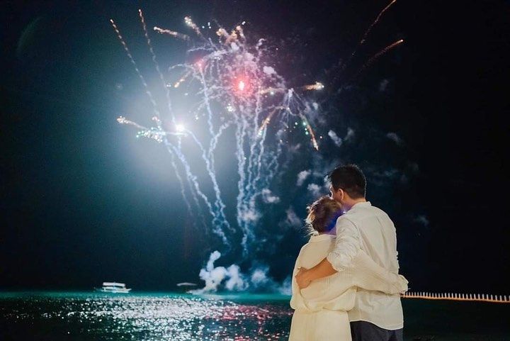 A bride and groom are kissing while watching fireworks over the ocean.