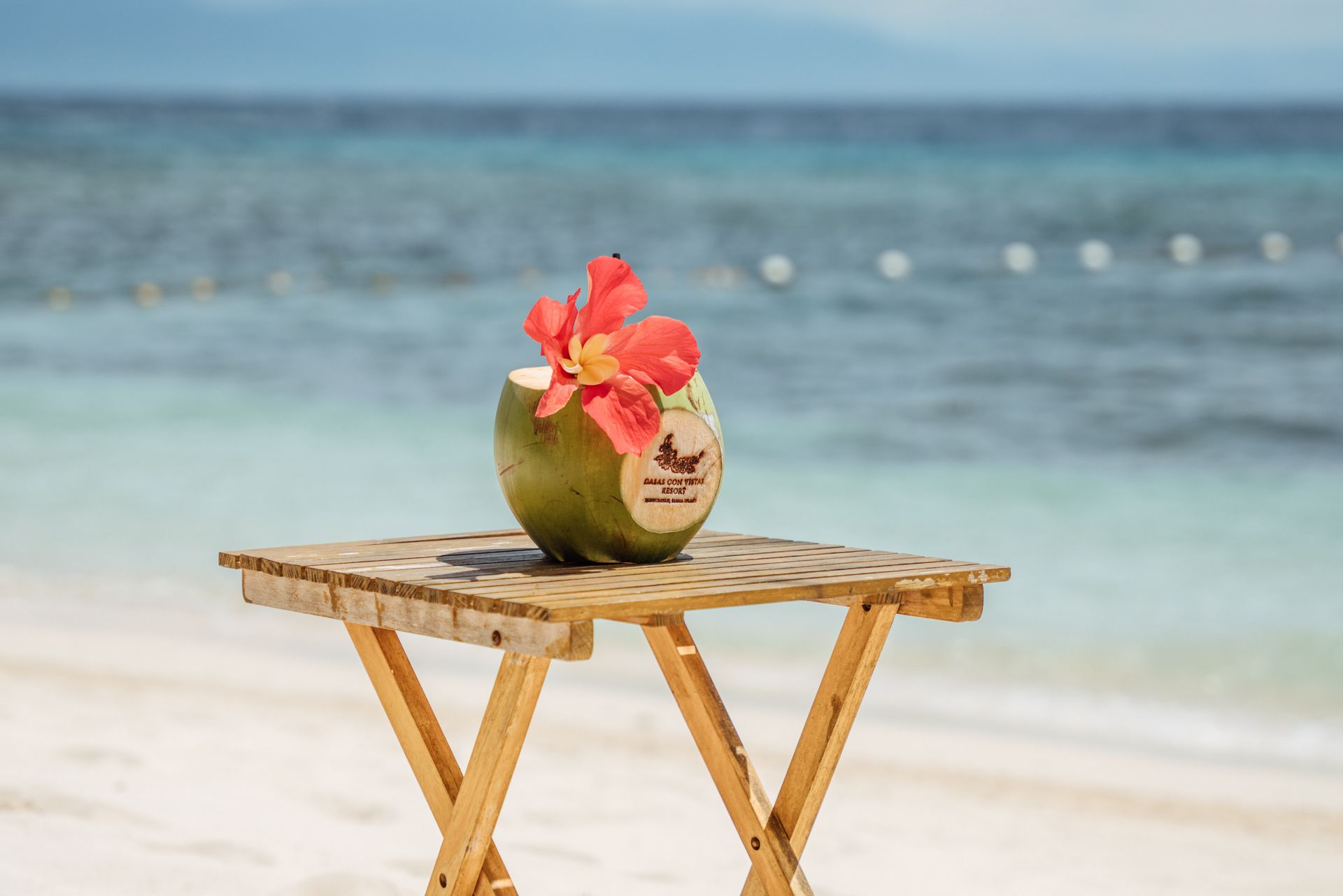 A coconut with a flower in it is on a wooden table on the beach.