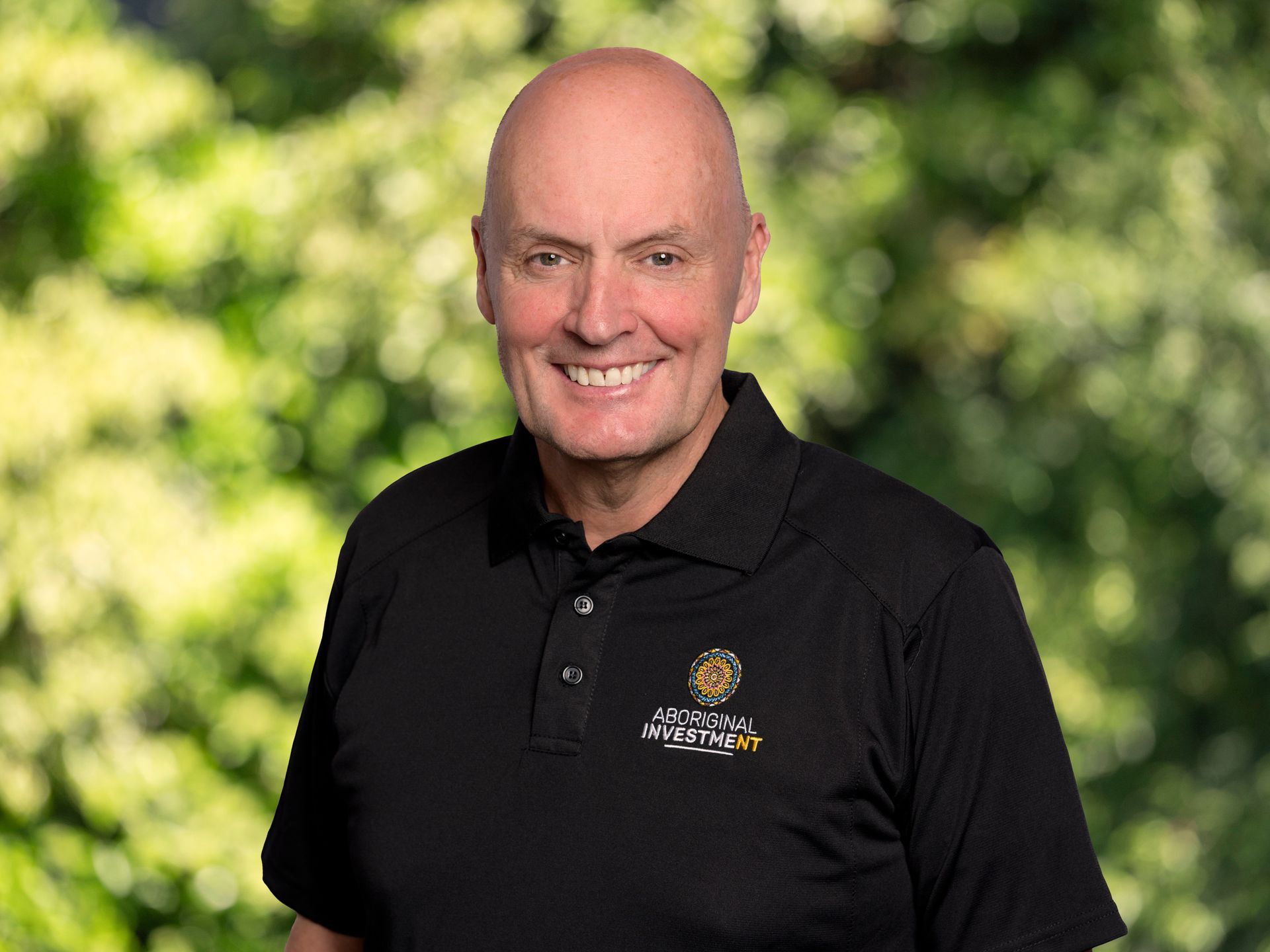 A bald man wearing a black polo shirt is smiling for the camera.