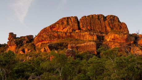 A large rocky mountain with trees in the foreground and a blue sky in the background.