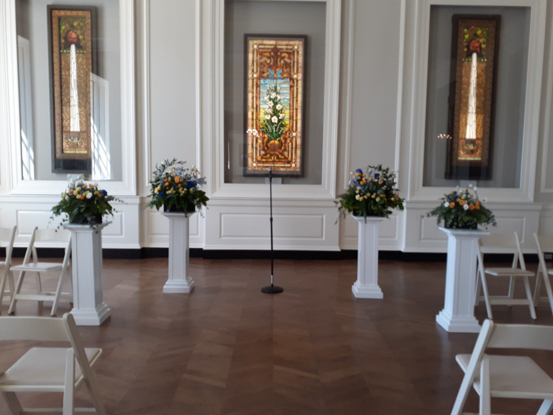 A room with a microphone and flowers on pedestals in front of a stained glass window.
