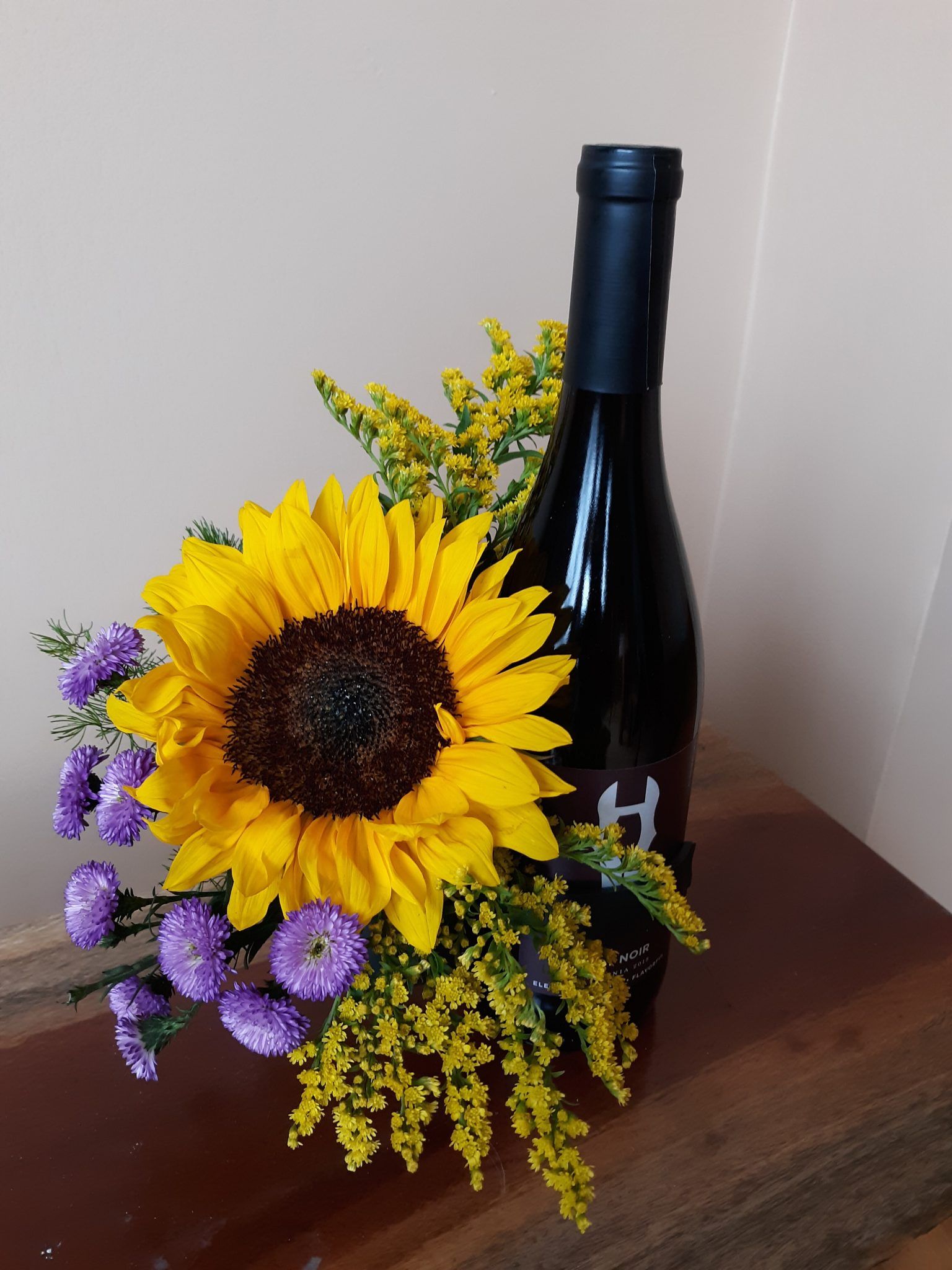 A bottle of wine with a sunflower and purple flowers in it