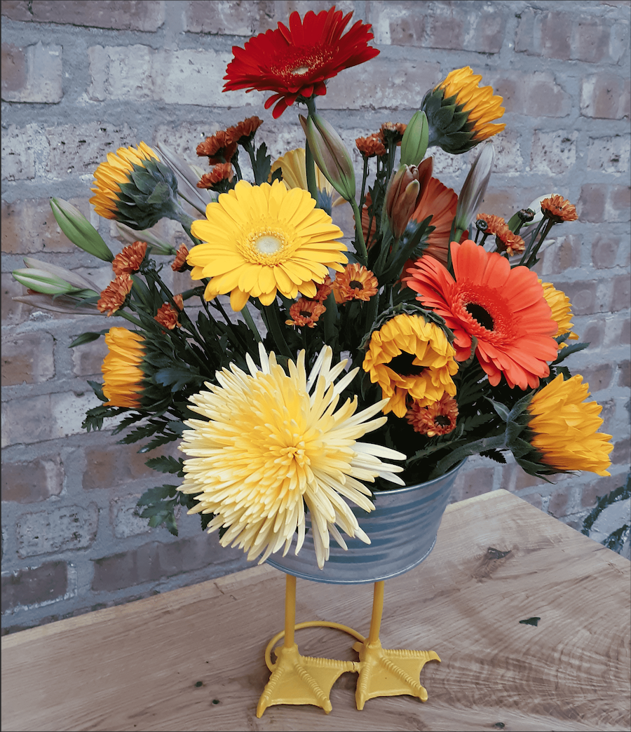 A bucket filled with yellow and orange flowers sits on a wooden table.