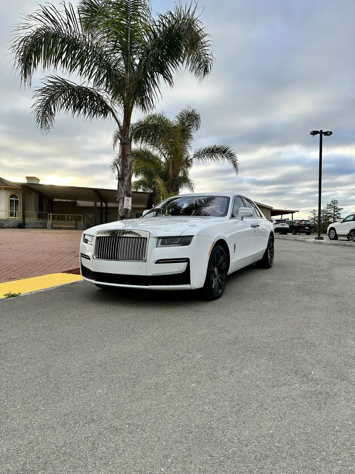 A white rolls royce ghost is parked in a parking lot next to a palm tree.