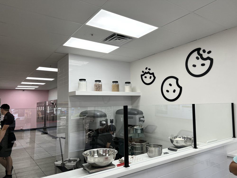 Cookie shop interior with cookie art on white wall, glass display, baking equipment, and customer.