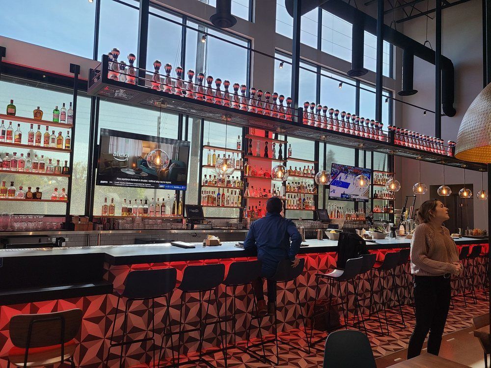 Interior view of a bar with patrons, bottles displayed, and TV screens, with red accents.