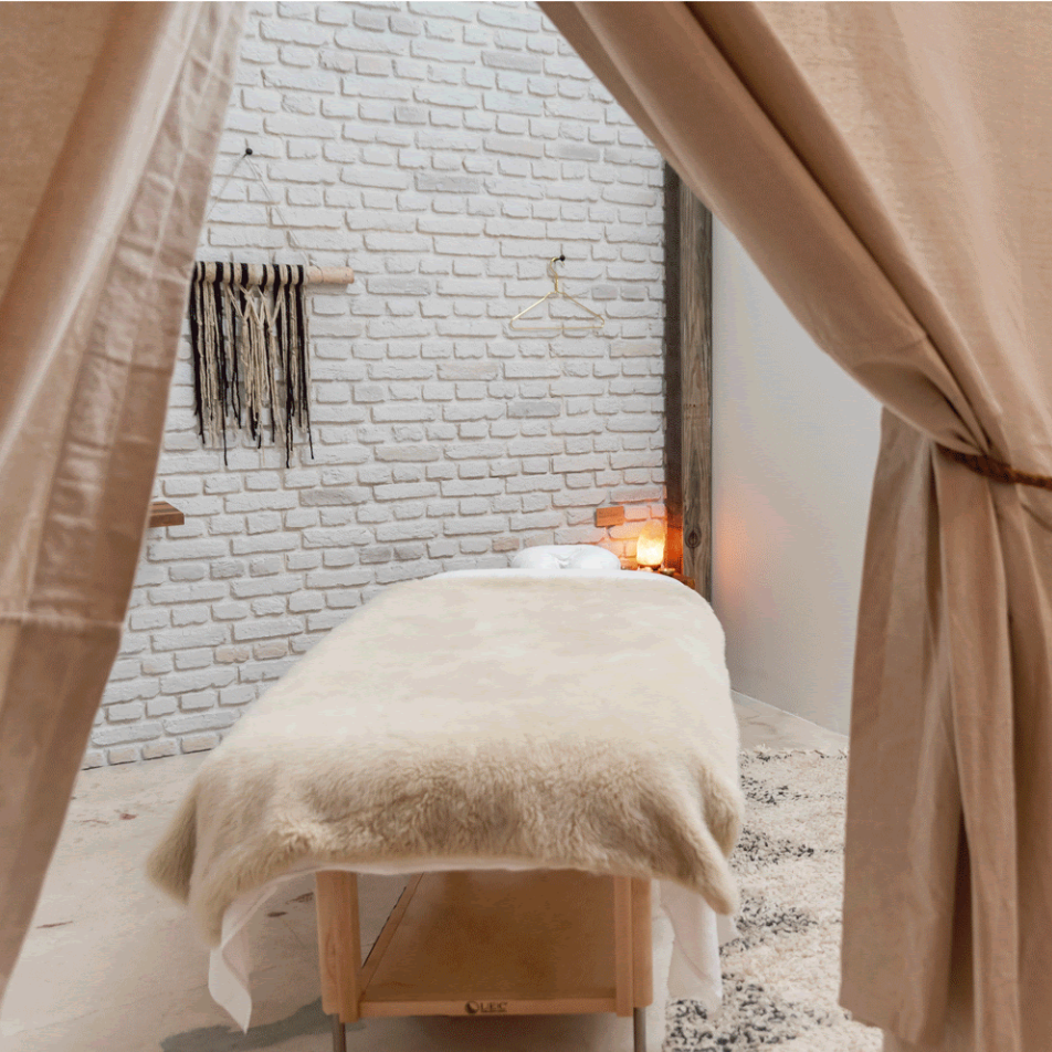 Massage table in a room with brick wall, lit candle, and curtains. Beige and white color scheme.
