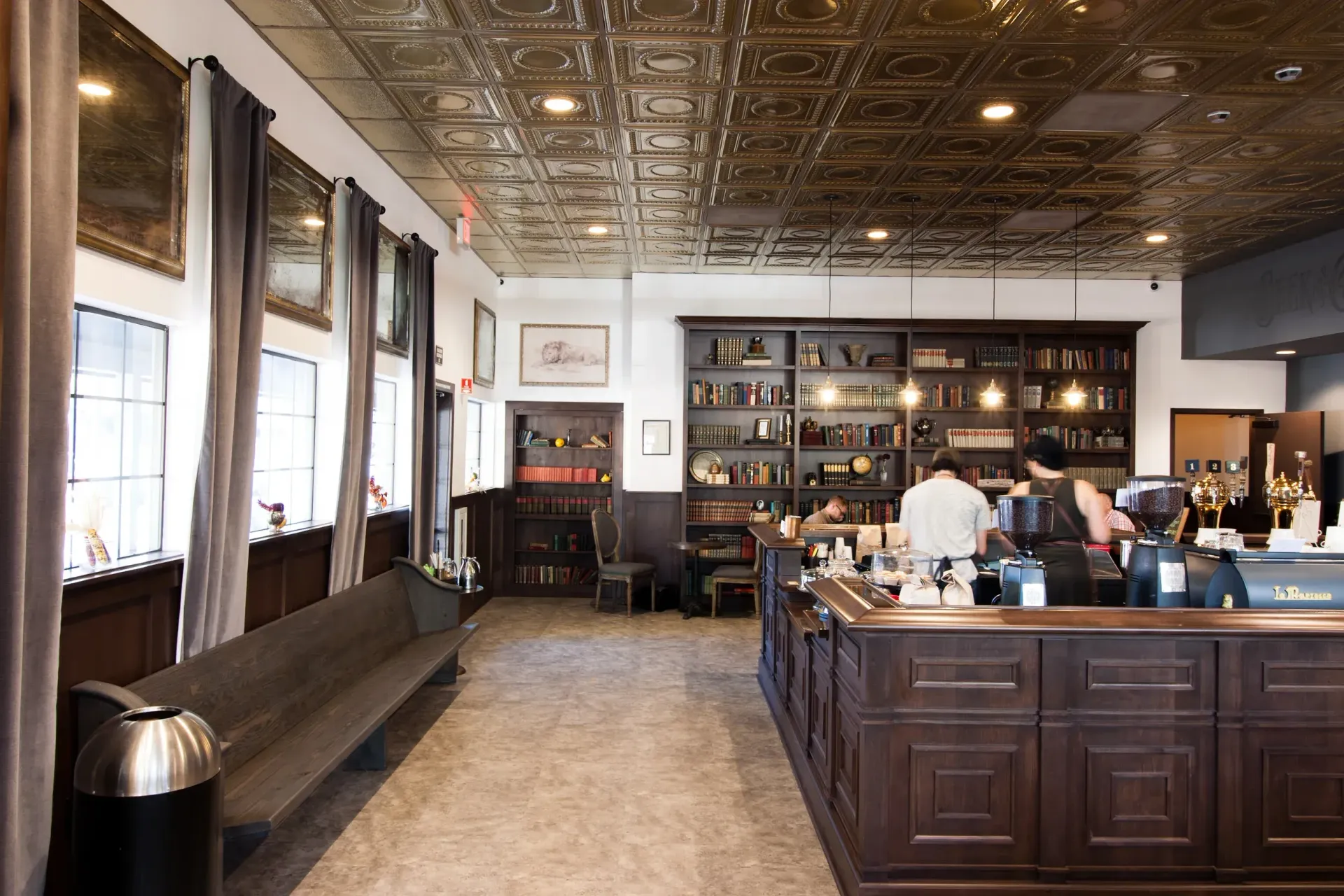 Inside a shop with dark wood counter and shelves, people are shopping. A long, dark wooden bench sits along the wall.