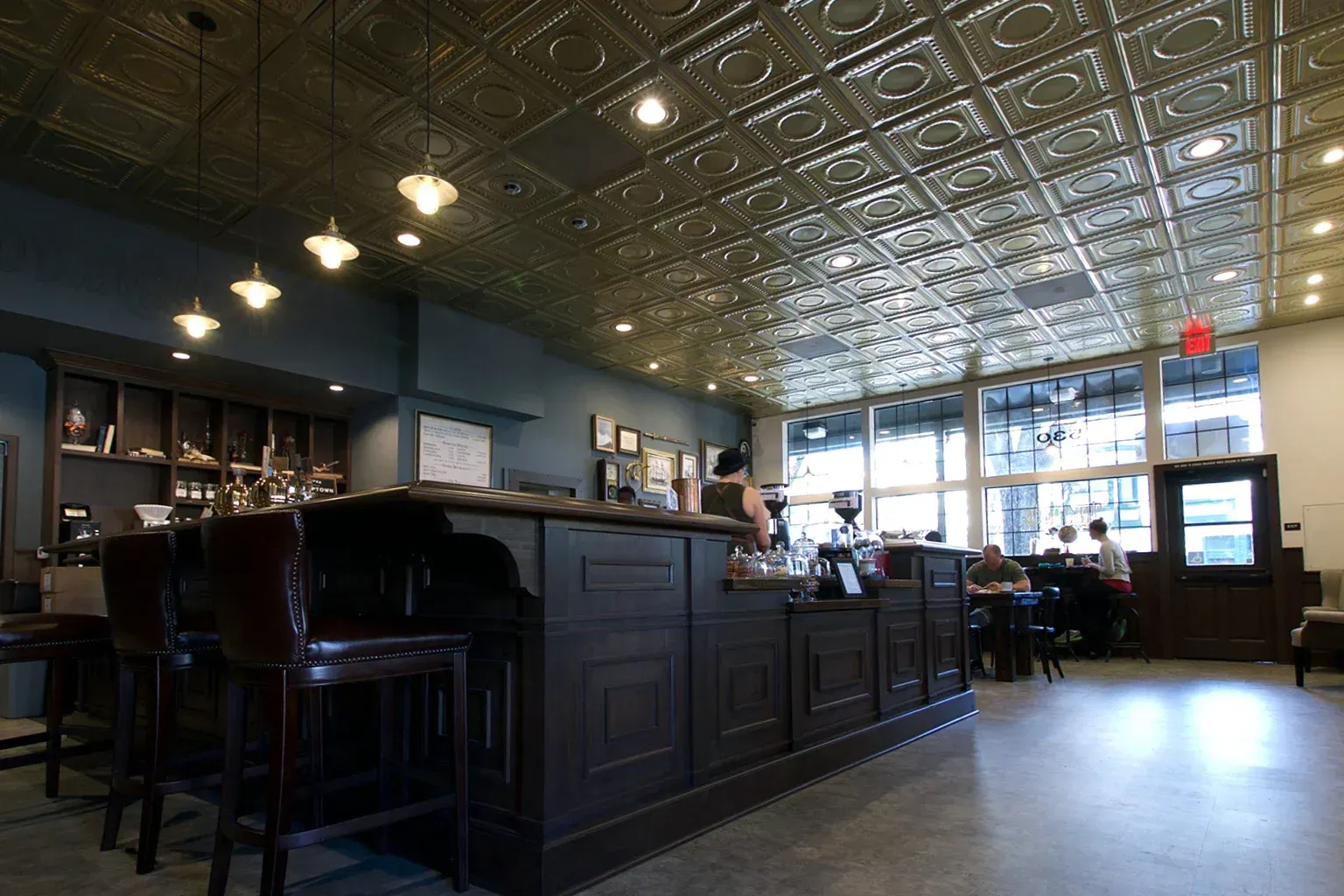 Coffee shop interior with dark wooden bar, patterned ceiling, and large windows.