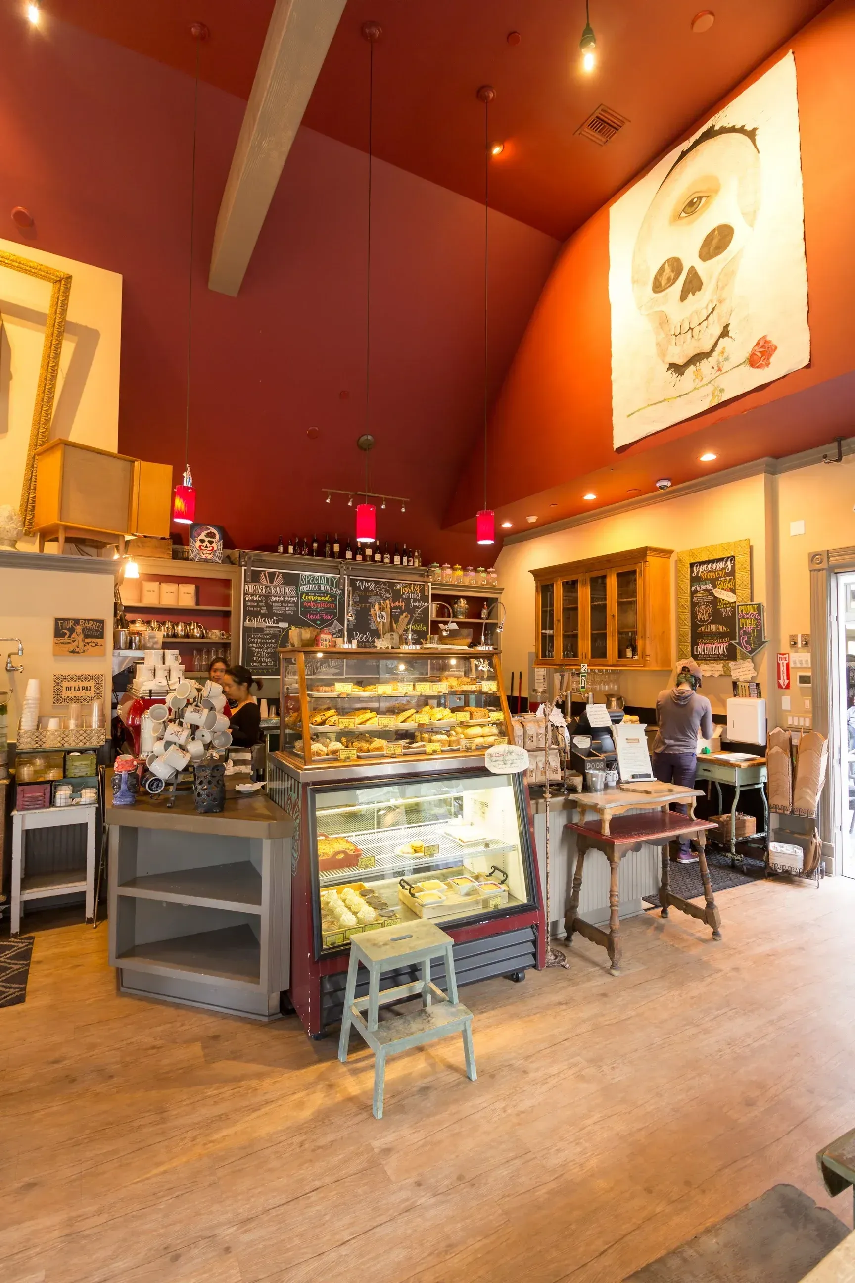 Cafe interior with baked goods display, artwork, and patrons. Red ceiling, wooden floors.