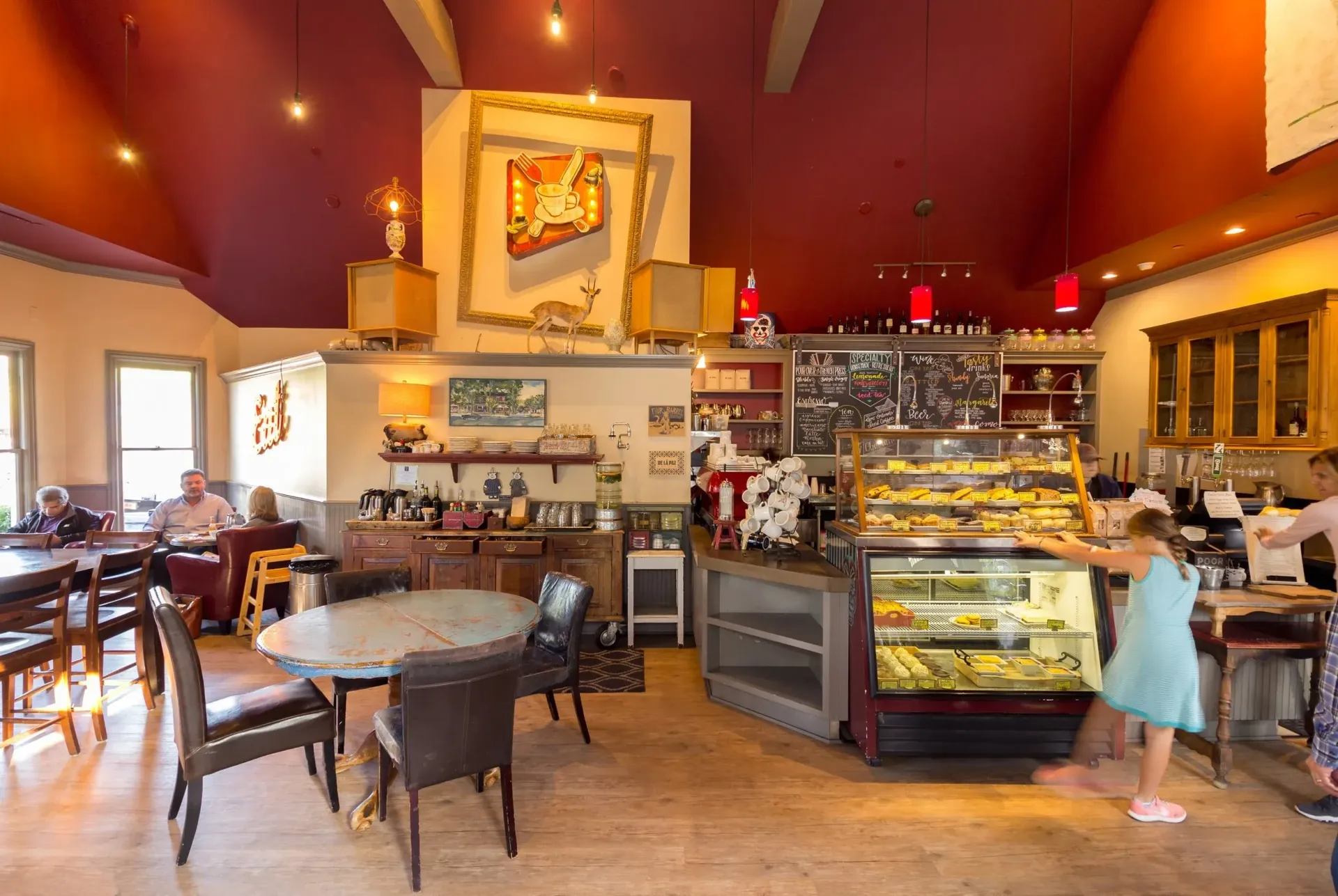 Interior of a cafe with tables, display case, and patrons. Red ceiling, wooden floors, bright lighting.