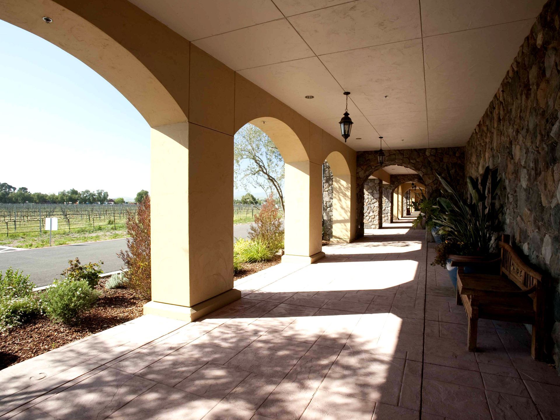 Covered walkway with arched openings, stone wall, and a view of vineyards.