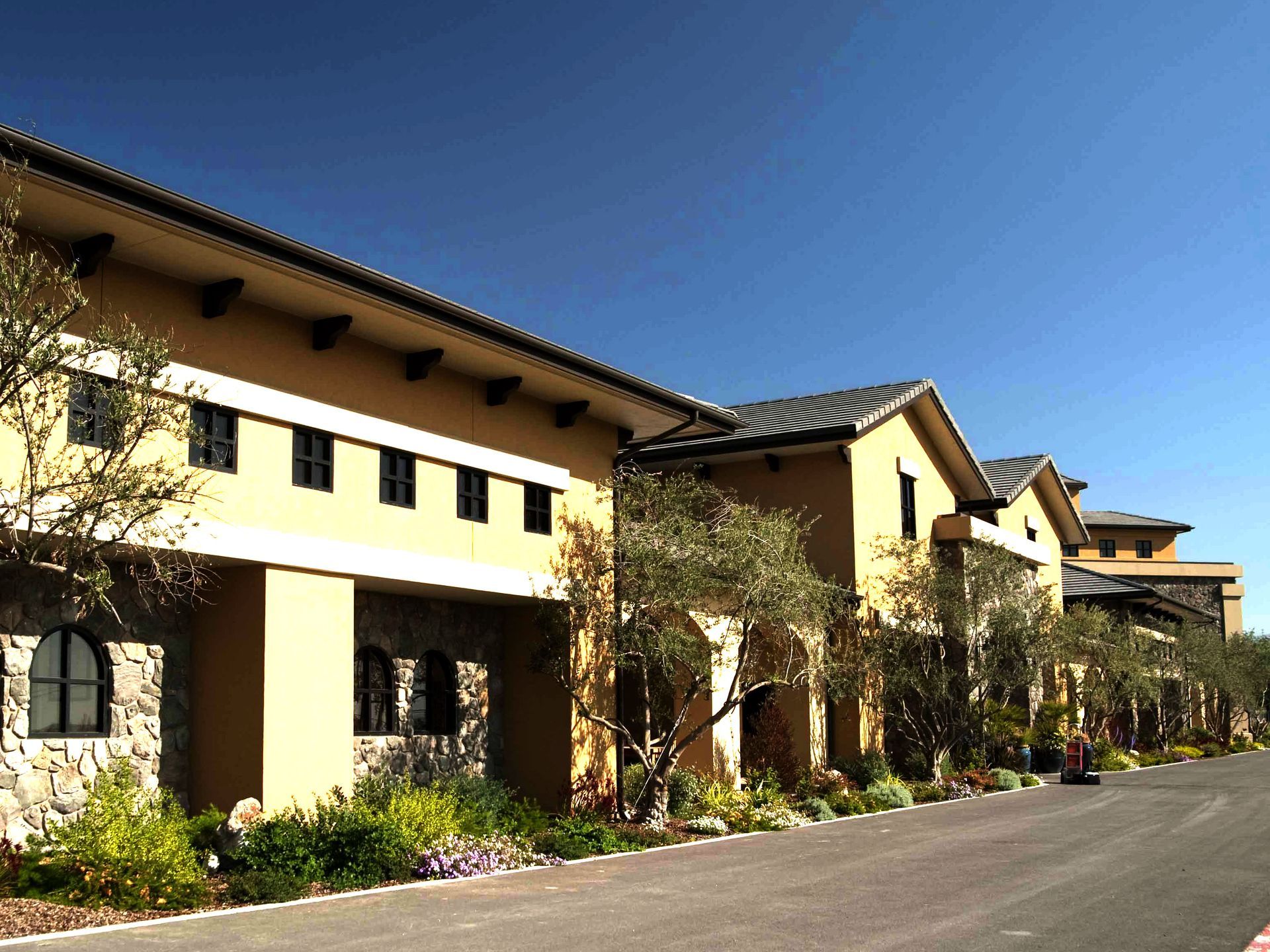 Beige stucco building with dark windows, tile roof, and olive trees under a blue sky.