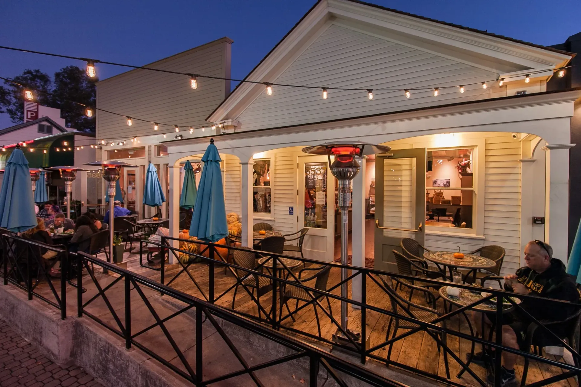 Outdoor restaurant patio at dusk, string lights, blue umbrellas, people dining, metal railings.