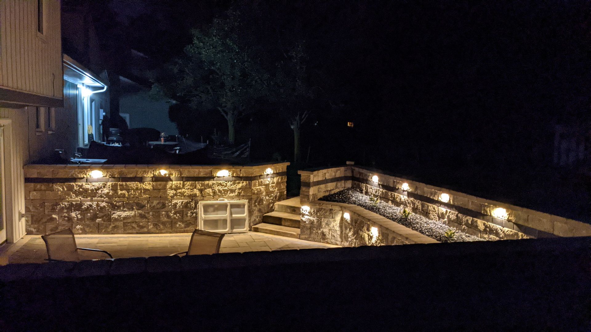 A patio with a stone wall and chairs is lit up at night.