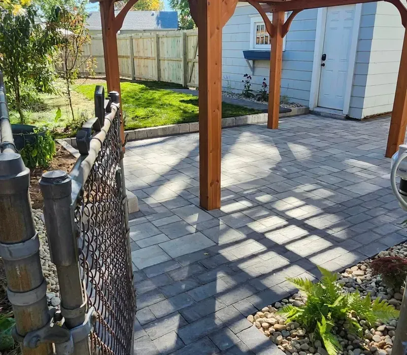 A wooden pergola is sitting on top of a brick patio next to a chain link fence.