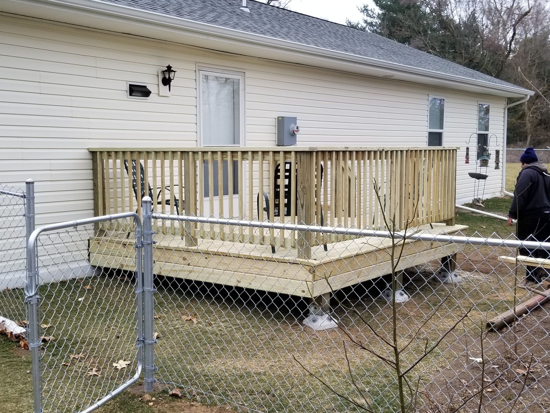 A man is standing in front of a house with a wooden deck behind a chain link fence.