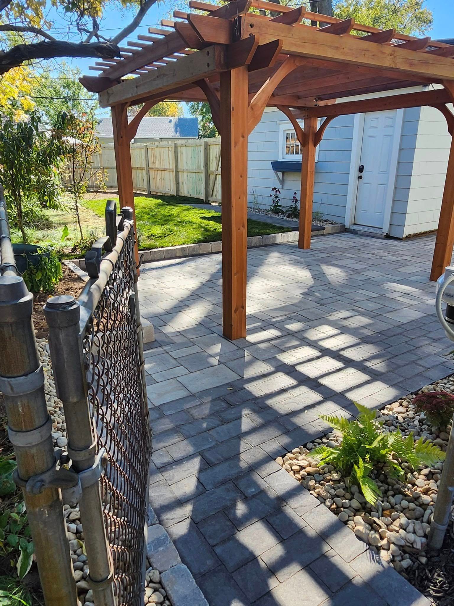 A wooden pergola is sitting on top of a brick patio next to a chain link fence.