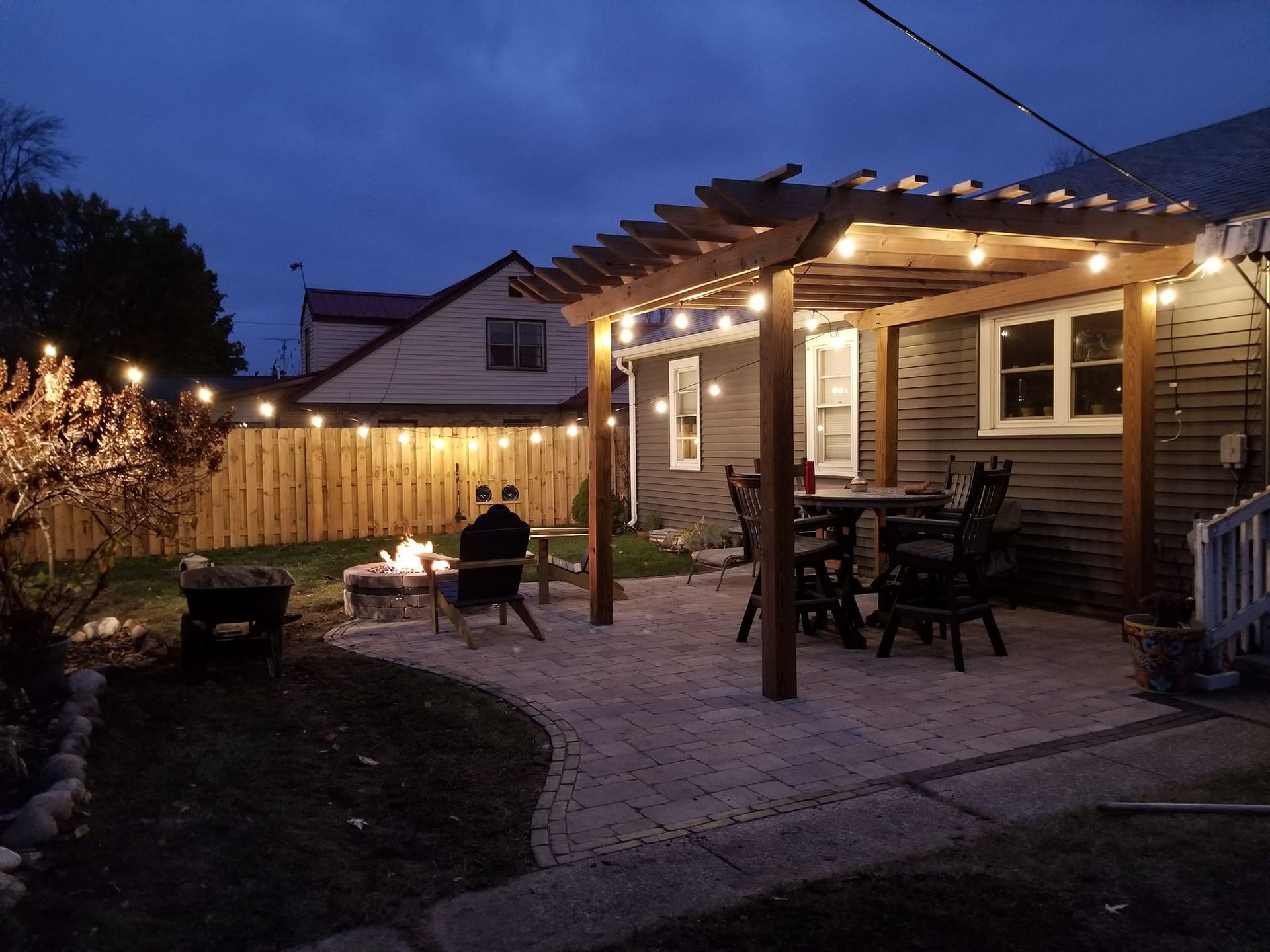 A backyard with a fire pit , table and chairs under a pergola at night.