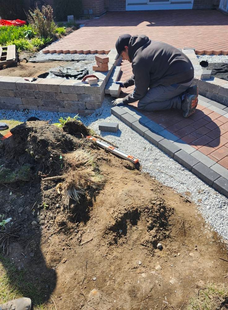 A man is kneeling down in the dirt while working on a brick walkway.