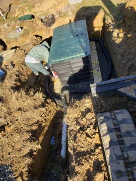 Worker crouches by stacked black trays in a dirt excavation site, handling equipment.
