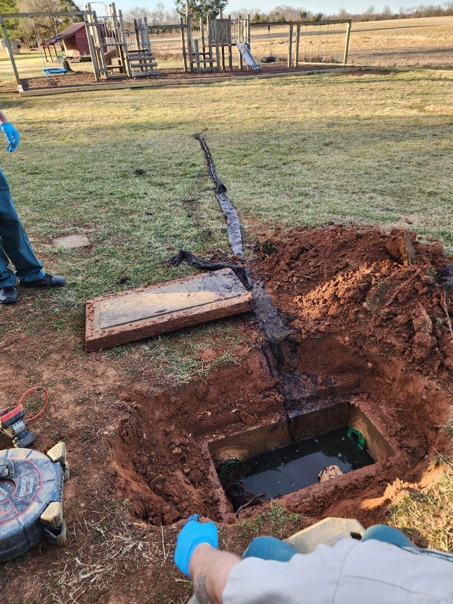 Open excavation trench in a yard with exposed pipe, soil pile, and workers’ gloved hands nearby