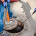 Worker opens manhole with a pry bar; black cable and drain cleaning tool are visible.