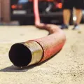 Close-up of a red fire hose with brass coupling lying on pavement, blurred firefighters in background