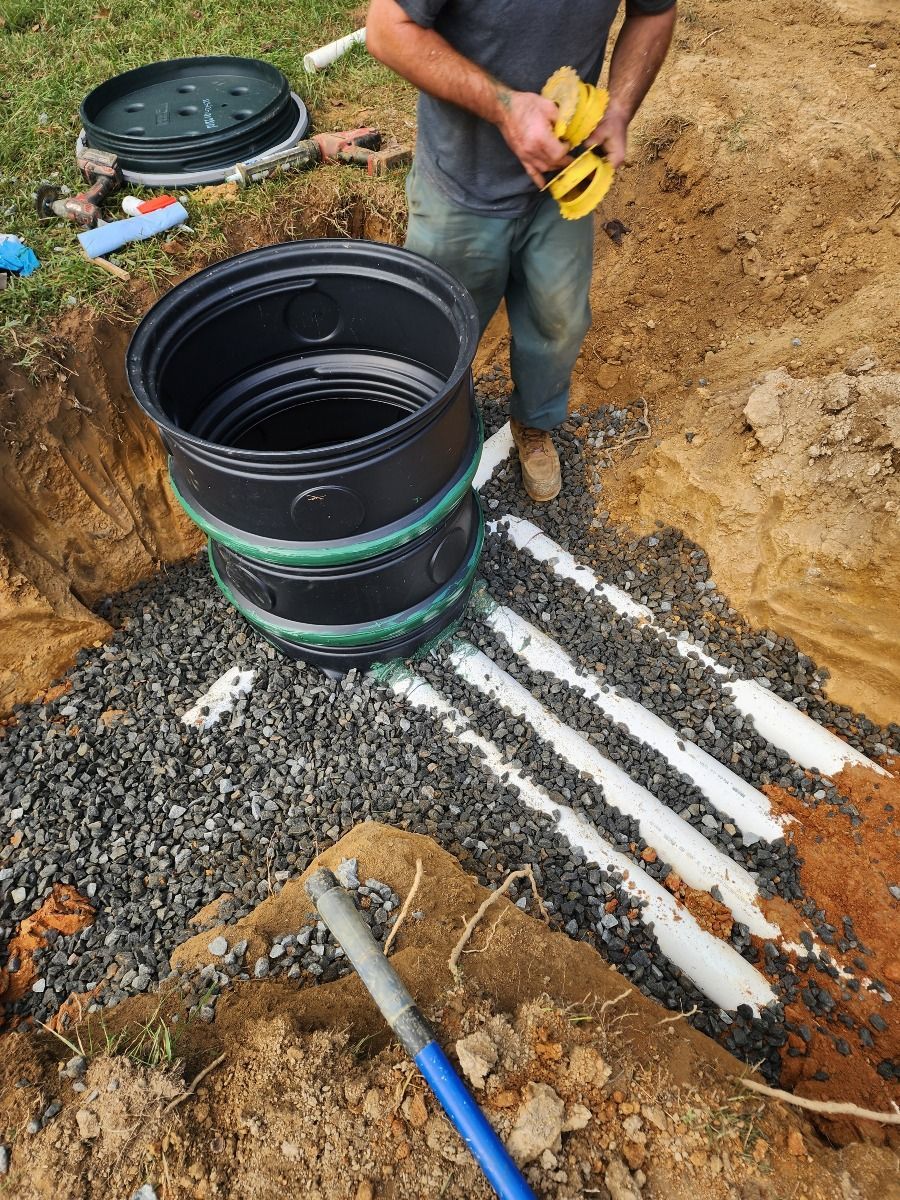 Worker installing a black plastic utility tank in a dirt trench beside white PVC pipes.