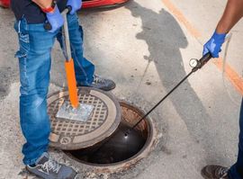 Workers inspecting a manhole with a sewer camera and orange pry bar on a street