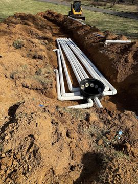 White PVC pipes laid in a dug trench at a construction site, with a mini excavator in the background.