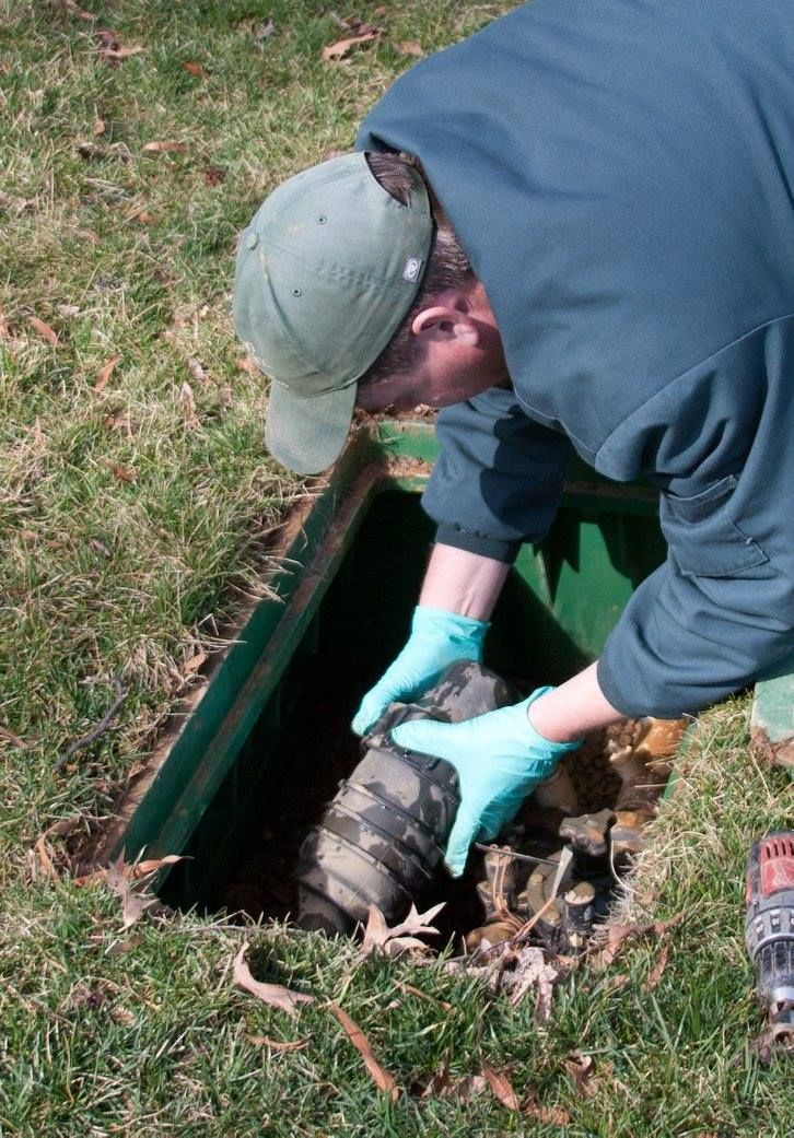 Person in a cap and gloves working at an open green utility hatch in the grass.