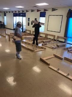 Two people inside a room assembling wooden frames. Man carrying wood, wearing a hard hat. Other man carrying wood on his shoulders.