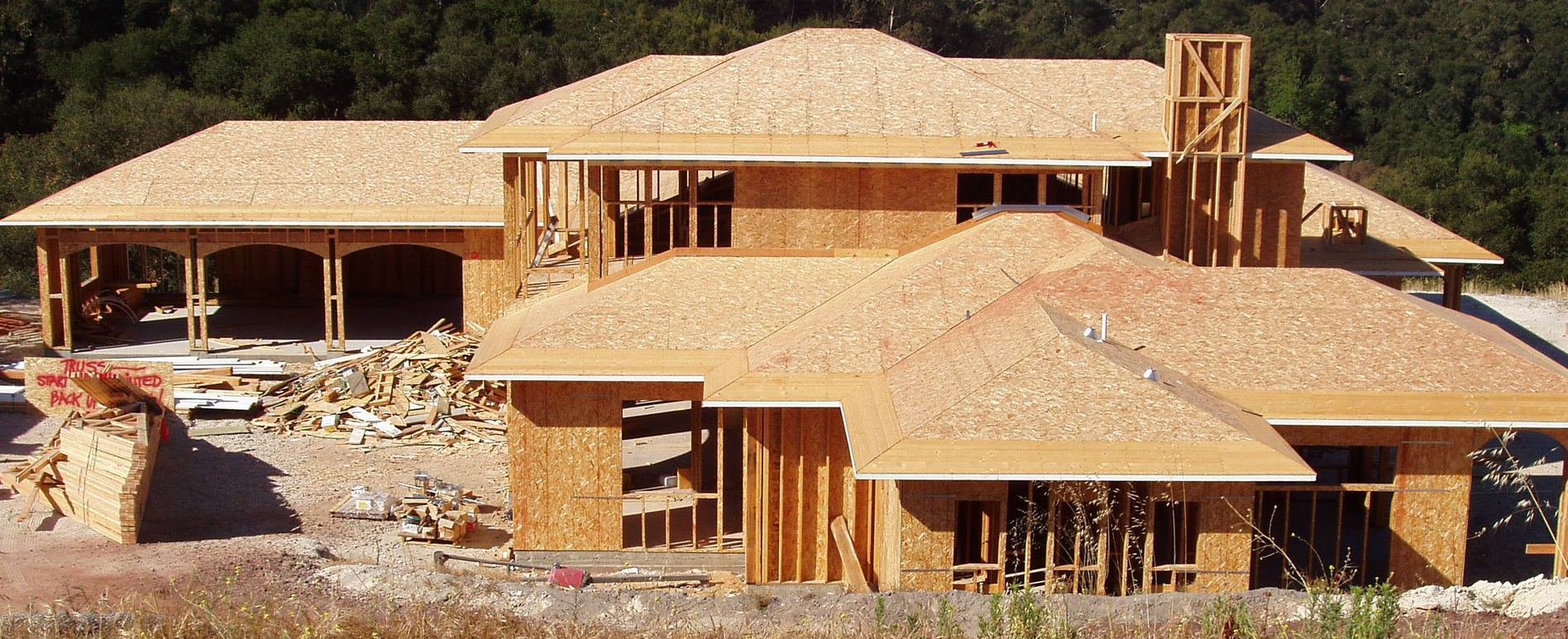 House under construction with wood framing and roofing, set against a backdrop of trees.